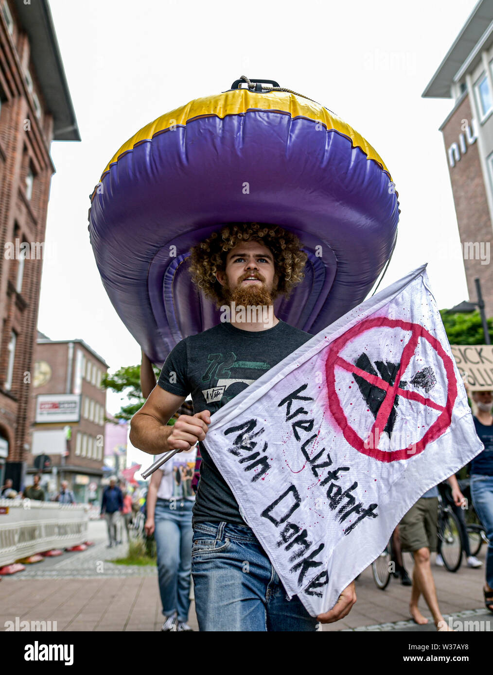 Kiel, Germany. 13th July, 2019. A participant of the demonstration 'Initiative gegen Kreuzfahrtschiffe' and 'TKKG' carries a rubber dinghy on his head and a poster with the inscription 'Kreuzfahrt Nein Danke' in his hands during the march through downtown Kiel. The ships 'MSC Meraviglia', 'Mein Schiff 1' and 'Aidaprima' dock in Kiel on 13 July. Credit: Axel Heimken/dpa/Alamy Live News Stock Photo
