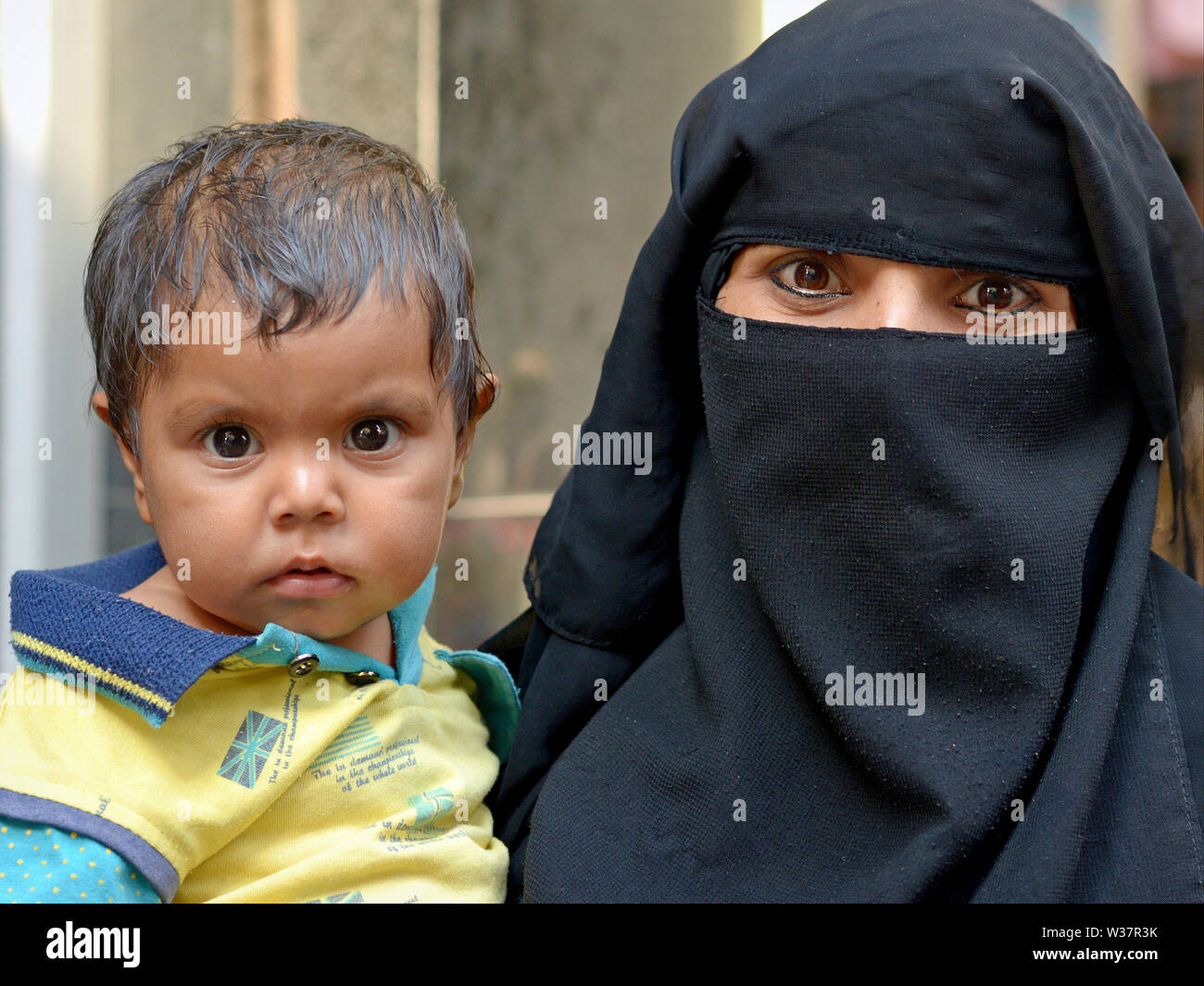 Indian Muslim woman with romantic eyes and eye makeup wears a black niqab and burqa and carries her toddler son in her arms. Stock Photo