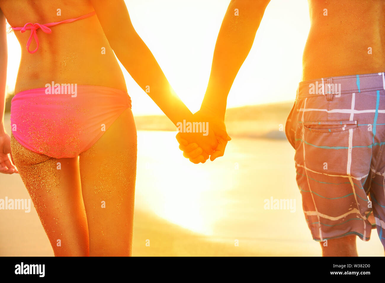 Holding hands couple in swimwear at beach. Rear view of fit couple's buttocks and legs as weight loss concept at beach sunset during summer vacations. Stock Photo