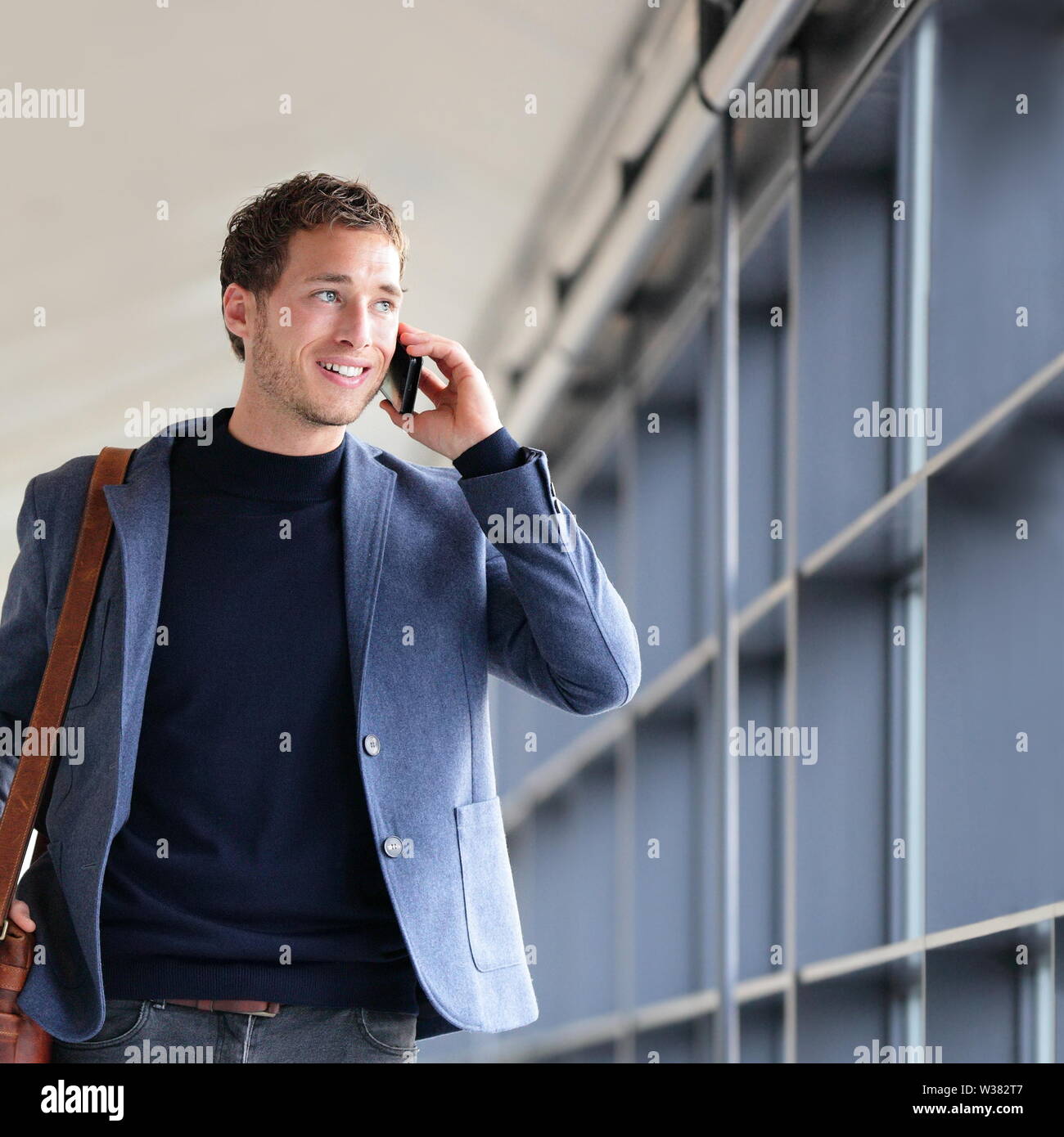 Urban business man talking on smart phone traveling walking in full body length inside in airport. Casual young businessman wearing suit jacket and shoulder bag. Handsome male model in his 20s. Stock Photo