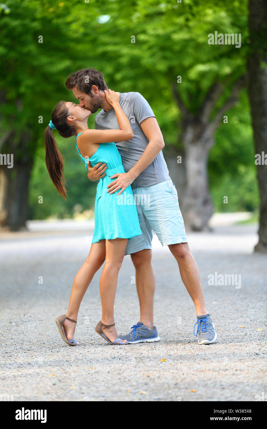 First kiss - Young couple of lovers in love passionately kissing standing  on path in summer park. Full body portrait of Caucasian male and Asian  female in blue sundress loving and hugging