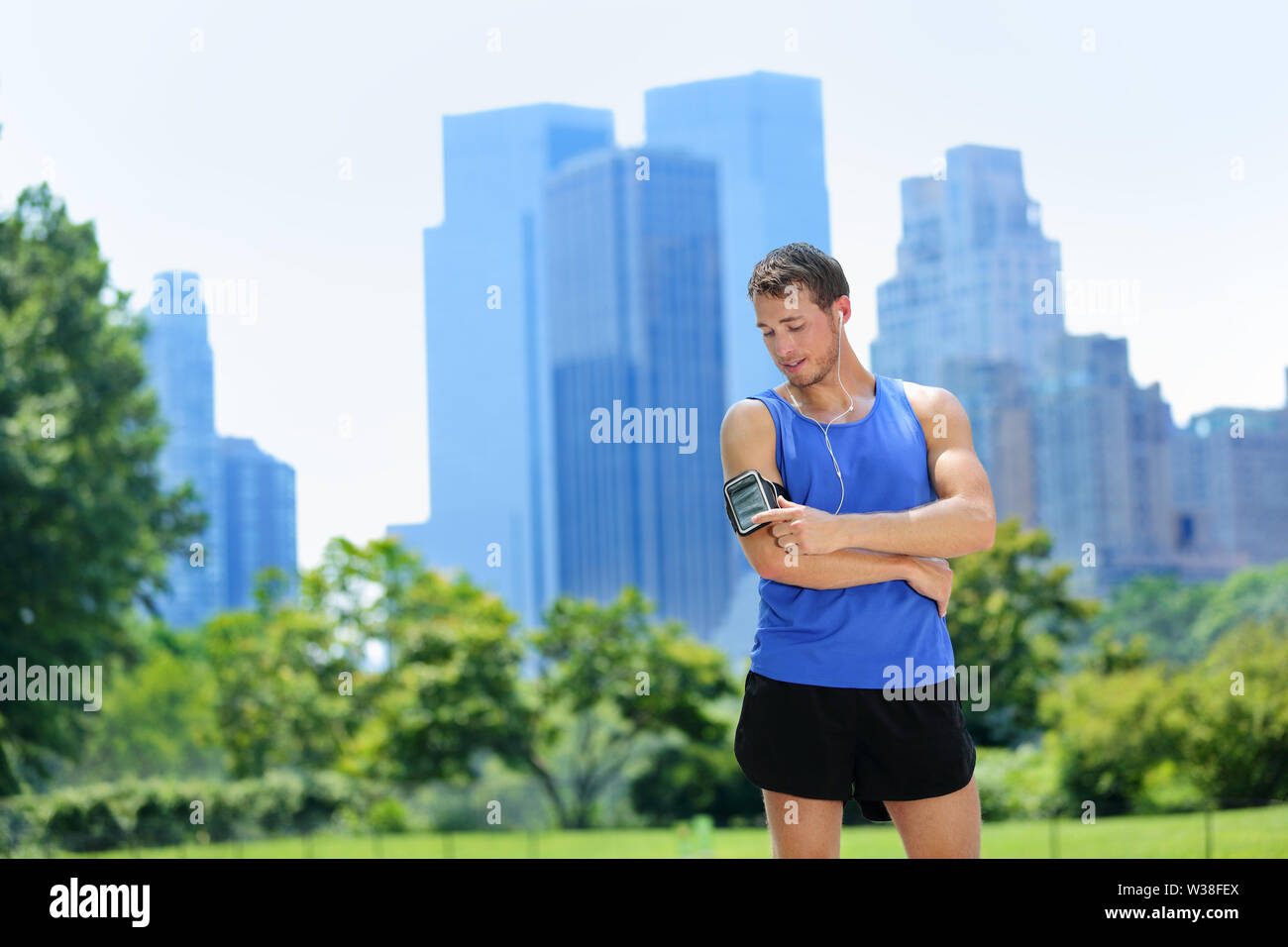 New York City man runner listening music on smartphone. Male adult jogger running using touchscreen on armband for workout in Central Park with urban background of Manhattan's skyscrapers skyline. Stock Photo