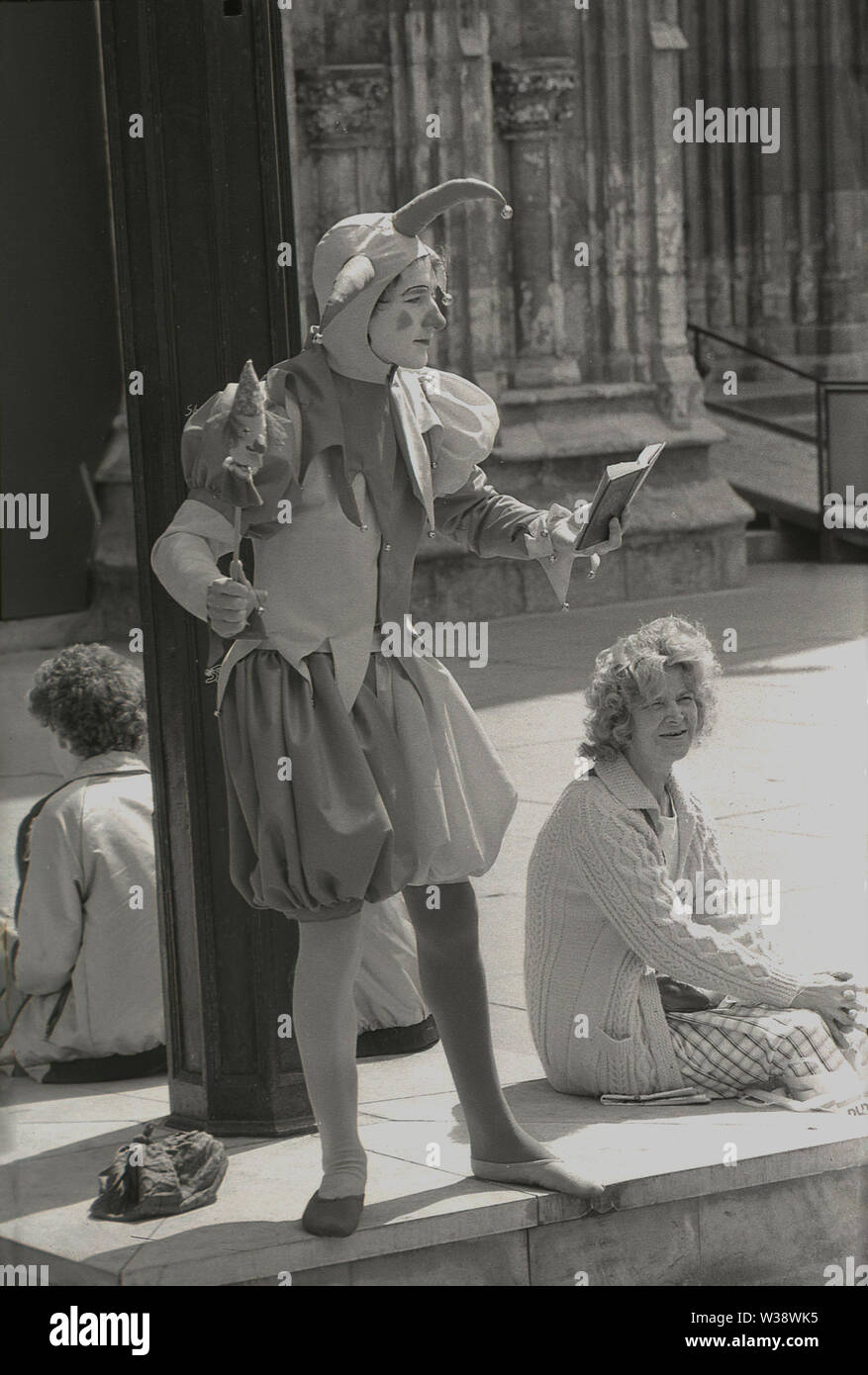 1980s, historical, a male jester, wearing traditional medieval costume, performing in the street, holding a marotte in one hand and reading a verse from a book, England, UK. Stock Photo