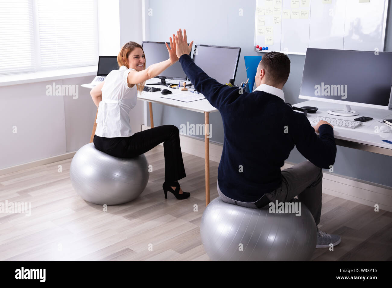 Businessman And Businesswoman Sitting On Fitness Ball Giving High Five In Office Stock Photo