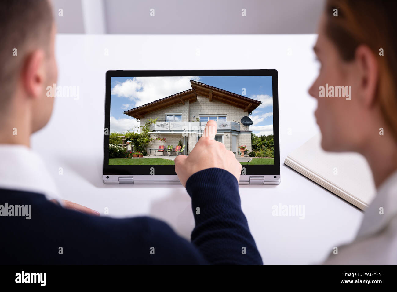 Male Estate Agent's Hand Pointing Laptop Screen Showing House Model Over Desk In Office Stock Photo