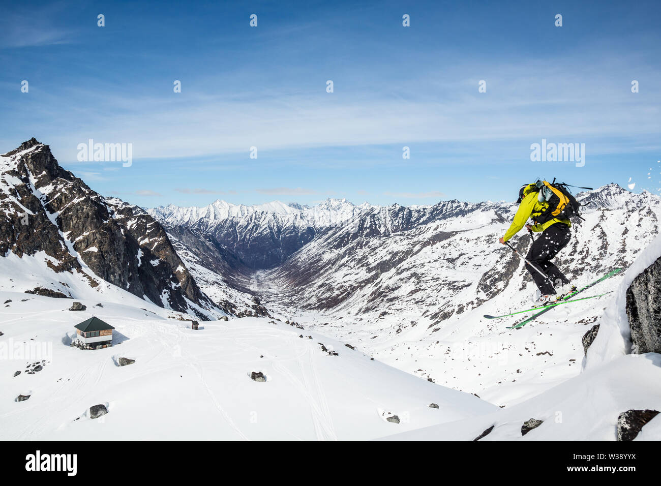 Skier above Snowbird hut and glacier jumping off a large boulder. Behind him is the Bartholf Creek drainage and a large couloir on the distant peak. Stock Photo