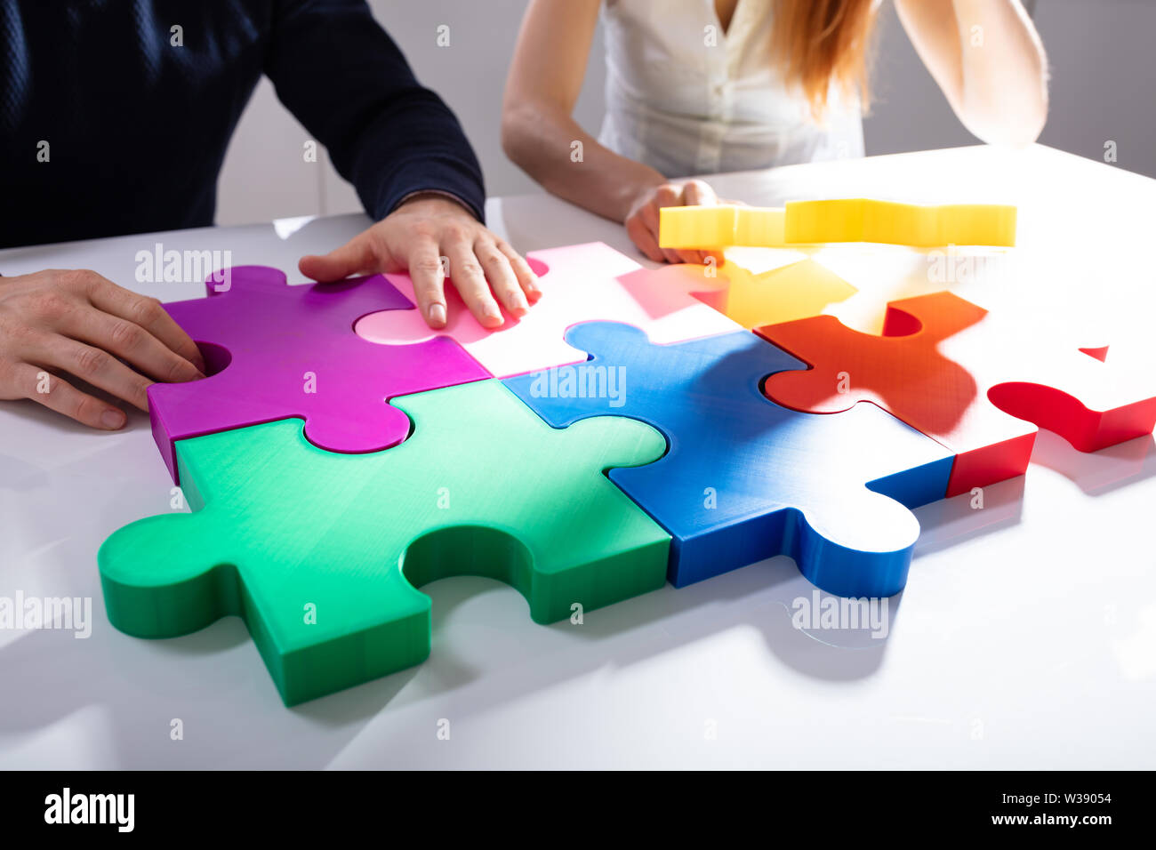 Two Businesspeople Solving Multi Colored Jigsaw Puzzle Over White Desk Stock Photo