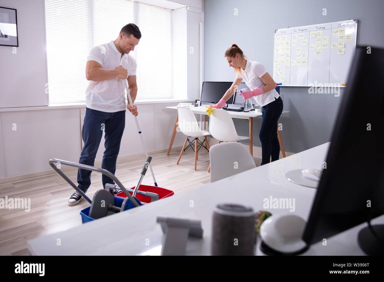 Two Smiling Young Janitor Cleaning The Desk And Mopping Floor In The Office Stock Photo