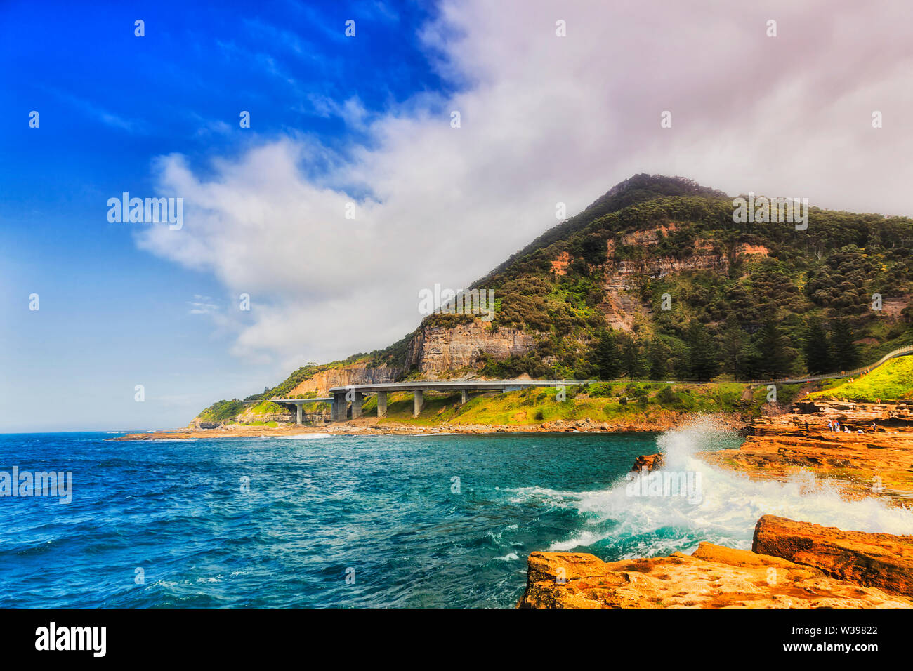 Sandstone rocks with surfing waves around Sea CLiff bridge of Grand Pacific drive in NSW, Australia, under blue sky. Stock Photo