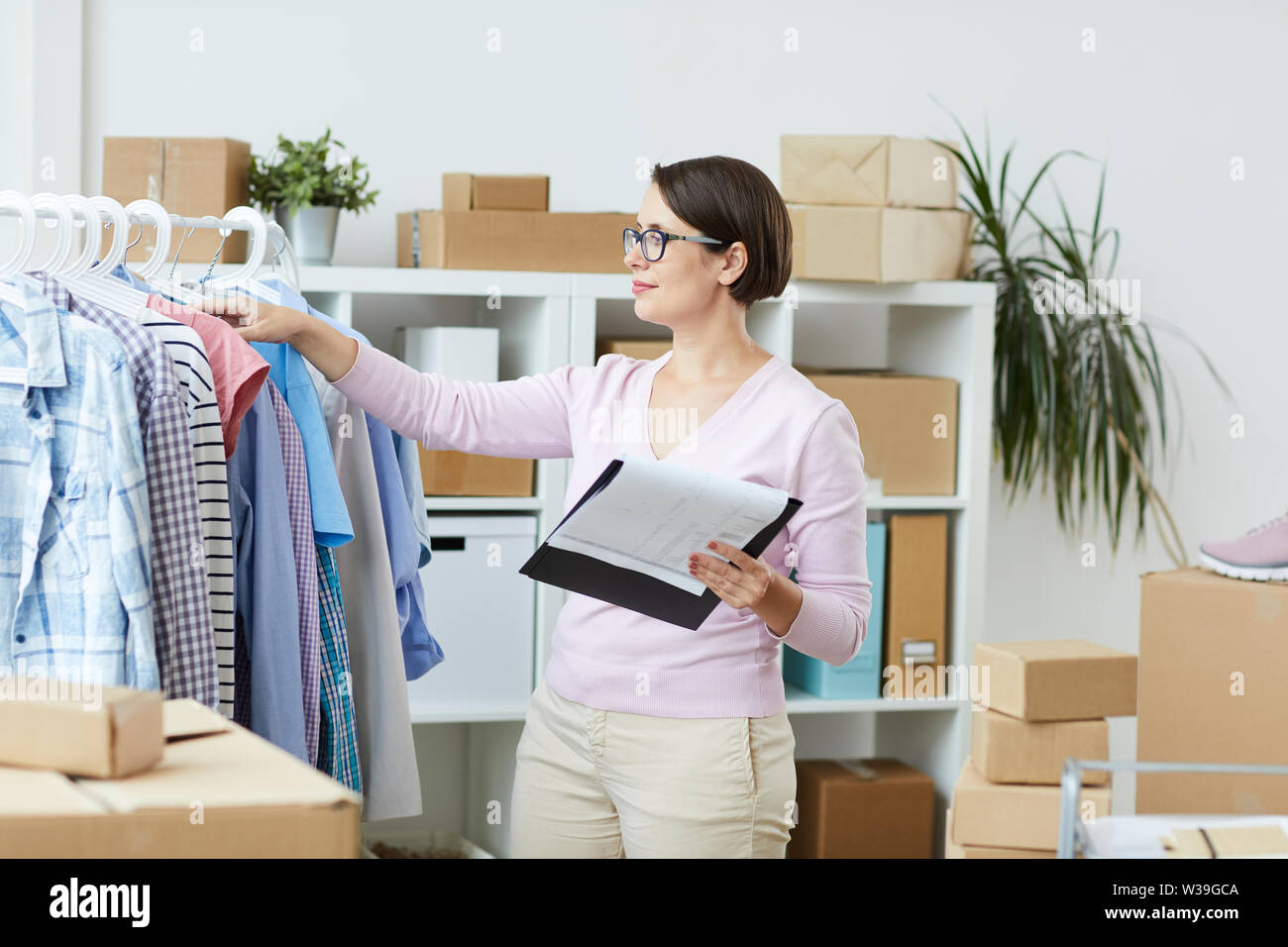 Young woman with document standing by rack and looking through new collection of casualwear Stock Photo
