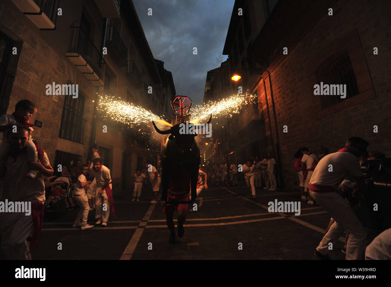 A man runs through the streets with a bull's head and horns that shoot out sparks in all directions during the San Fermin bull running fiesta in Pamplona. The 'Toro de Fuego' is a typical sight every night during the San Fermin fiesta where children have their particular confinement of an artifact made of cardboard carried by a young man. Stock Photo