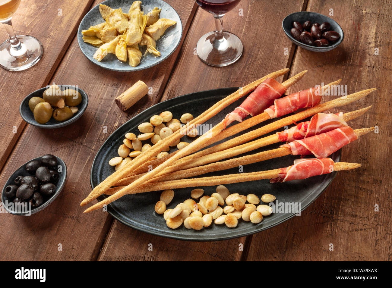 Italian antipasti. Grissini breadsticks with prosciutto di parma and roasted almonds, with olives and artichokes, with wine glasses Stock Photo
