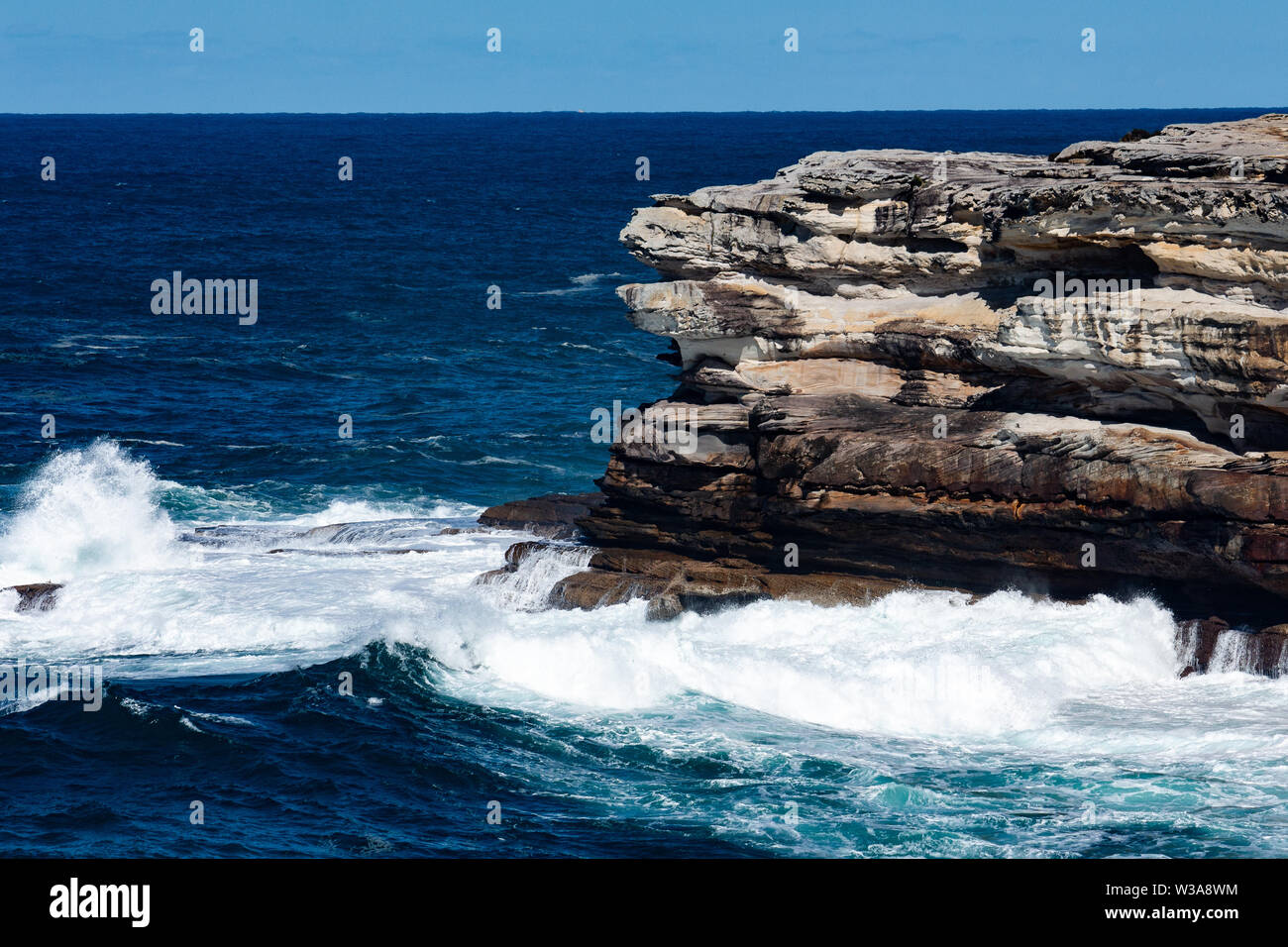 Oceanside rocky sandstone cliff with blue sea water waves creating whitewash against coastline and clear sky in background Stock Photo