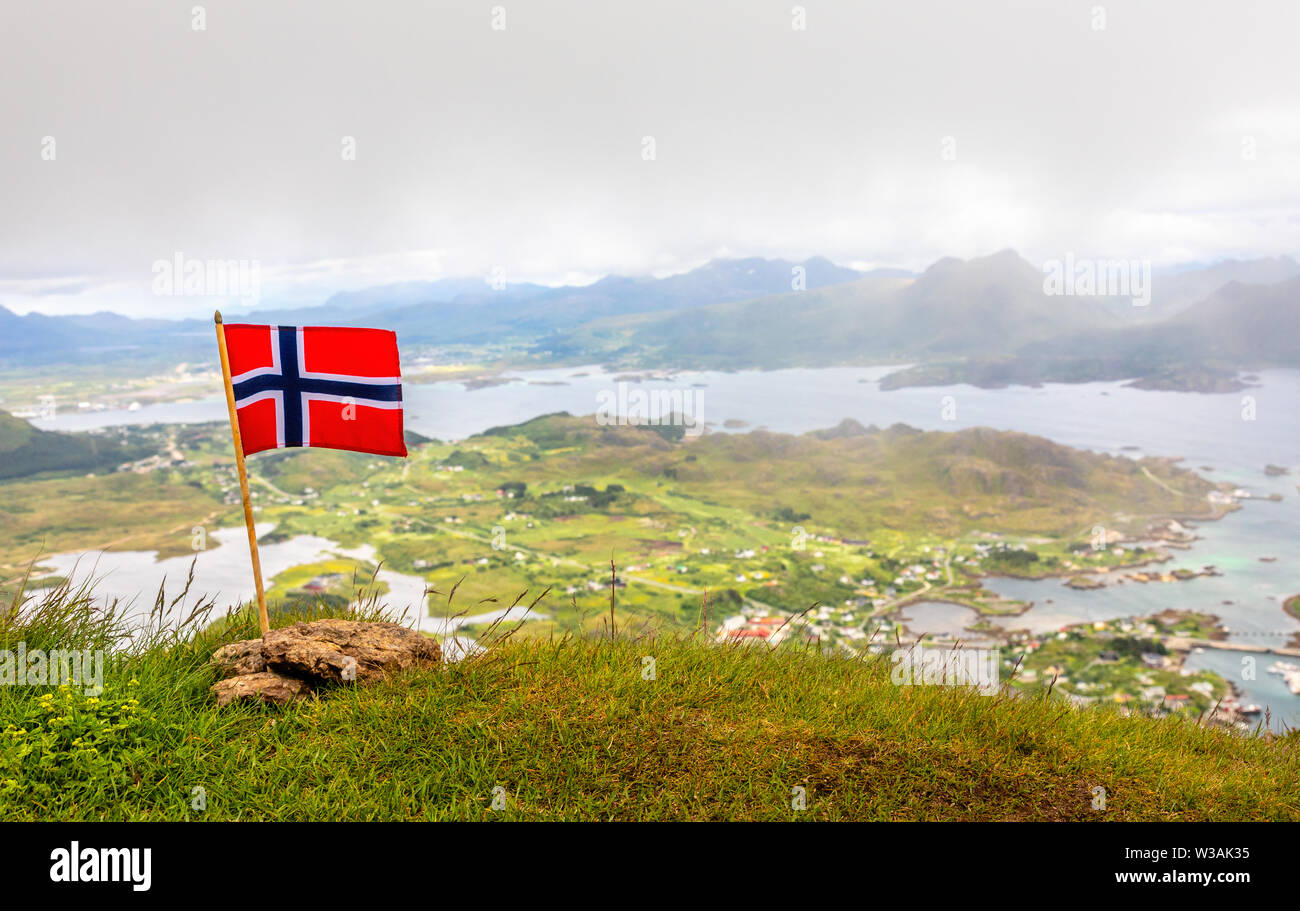 Norwegian national flag in the wind on the top of Nonstinden peak with fjord in the background, Ballstad, Vestvagoy Municipality, Nordland county, Nor Stock Photo