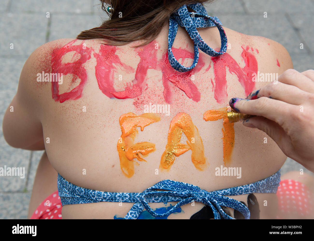 Berlin, Germany. 14th July, 2019. Bjale paints Alisa with the stroke "Burn  Fat" during the body painting at the protest action "2. Berlin Bikini- und  Badehose Bicycle Ride". The participants then demonstrated