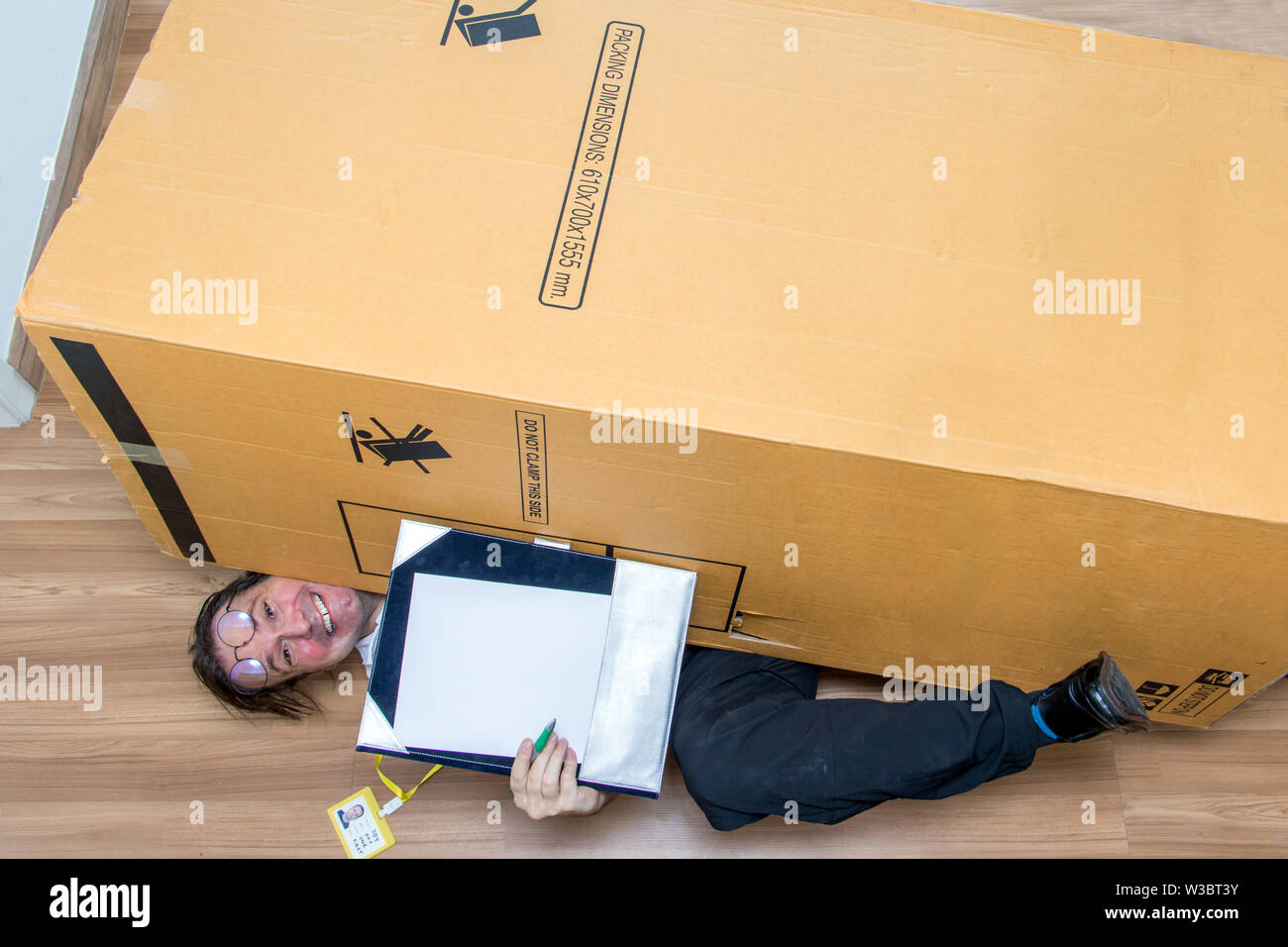The mailman lying under a big box and show paper document for confirm shipment. Large cardboard fall down at the postman. Stock Photo