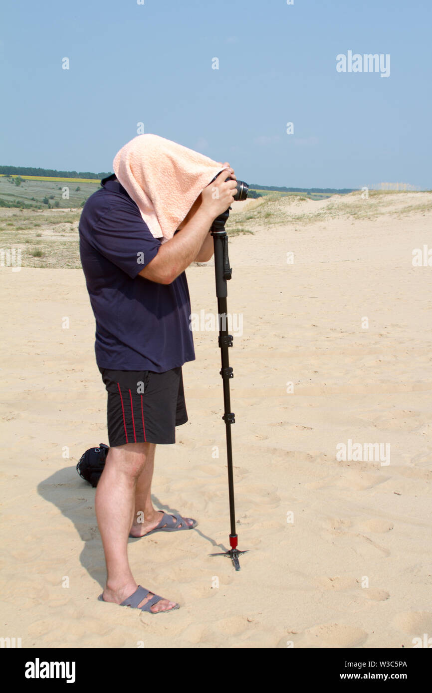 Kharkiv, Ukraine - July 31, 2016: Adult male photographer with a towel on his head from the heat is taking pictures with a camera on a stand in the de Stock Photo