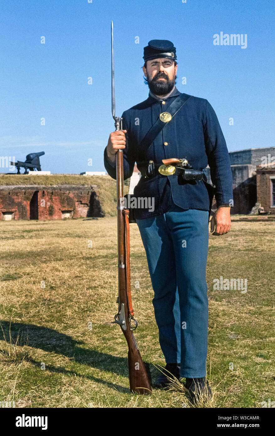 A bearded man wearing the military uniform of a U.S. Civil War Union soldier poses with weapons he carries as an interpreter who greets visitors to Fort Clinch State Park in Fernandina Beach on the east coast of Florida, USA. His right hand holds a Springfield rifled musket with its bayonet attached, and a pistol is tucked in the leather belt around his waist that also carries ammunition. This historic fort at the entrance to Cumberland Sound and the St. Mary’s River was initially occupied by the Confederate Army until it came under control of Union troops in 1862. Stock Photo