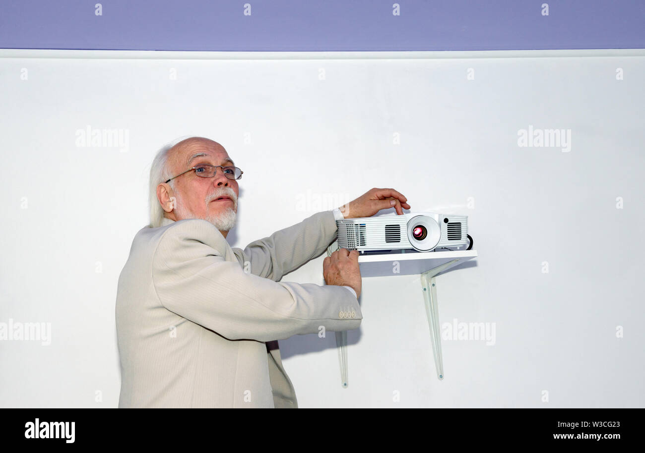 Portrait of senior grey bearded bald man in light business suit that is setting up the projector in office interior on white wall background. Stock Photo