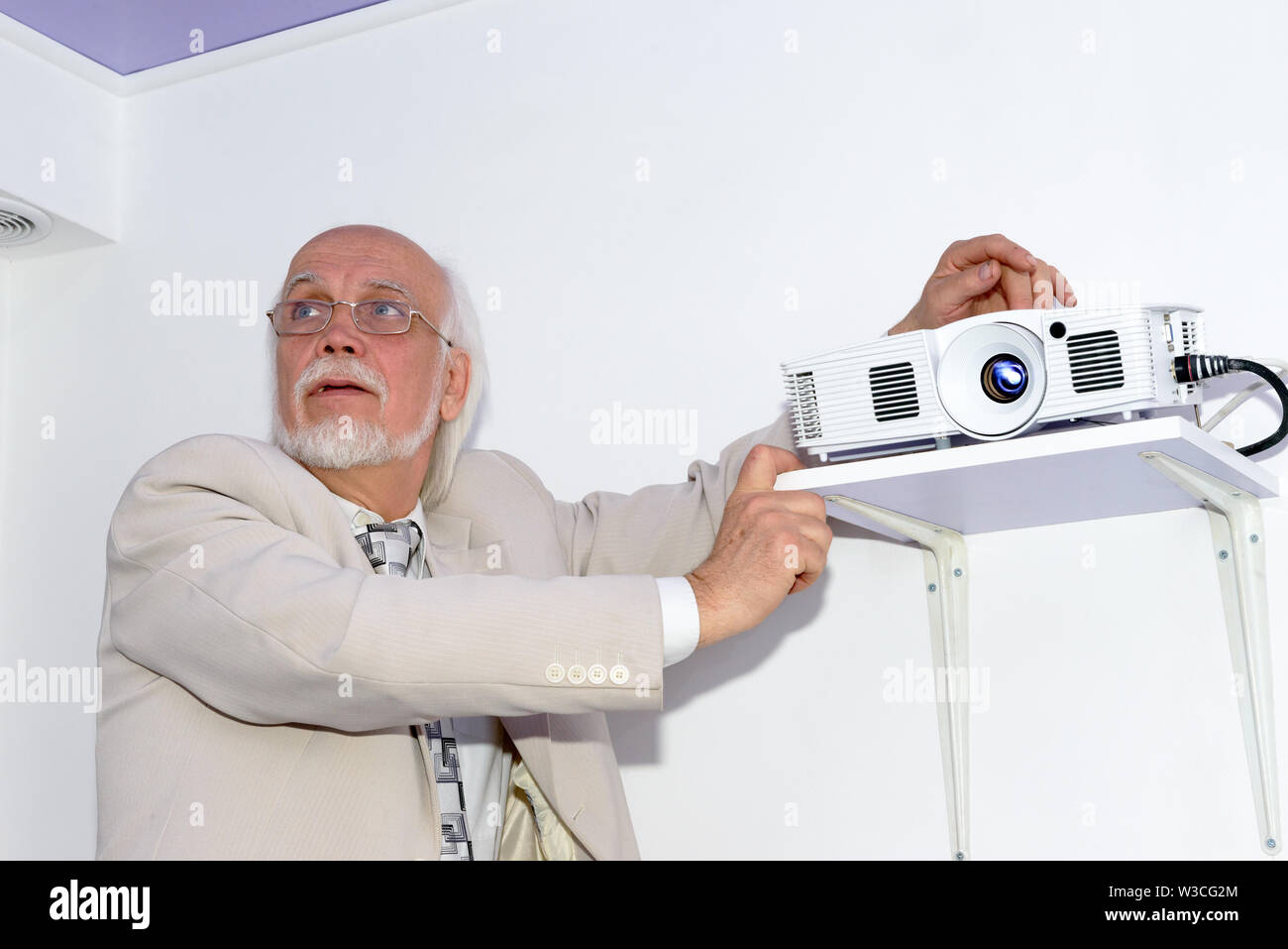 Portrait of senior grey bearded bald man in light business suit that is setting up the projector in office interior on white wall background. Stock Photo