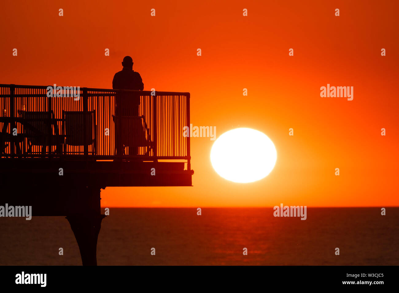 Aberystwyth Wales UK, Sunday 14 July 2019  UK Weather: People silhouetted against the orange colour of the sunset as they stand on  the seaside pier  in Aberystwyth at the end of a warm summers day  in west Wales.   photo credit: Keith Morris/Alamy Live News Stock Photo