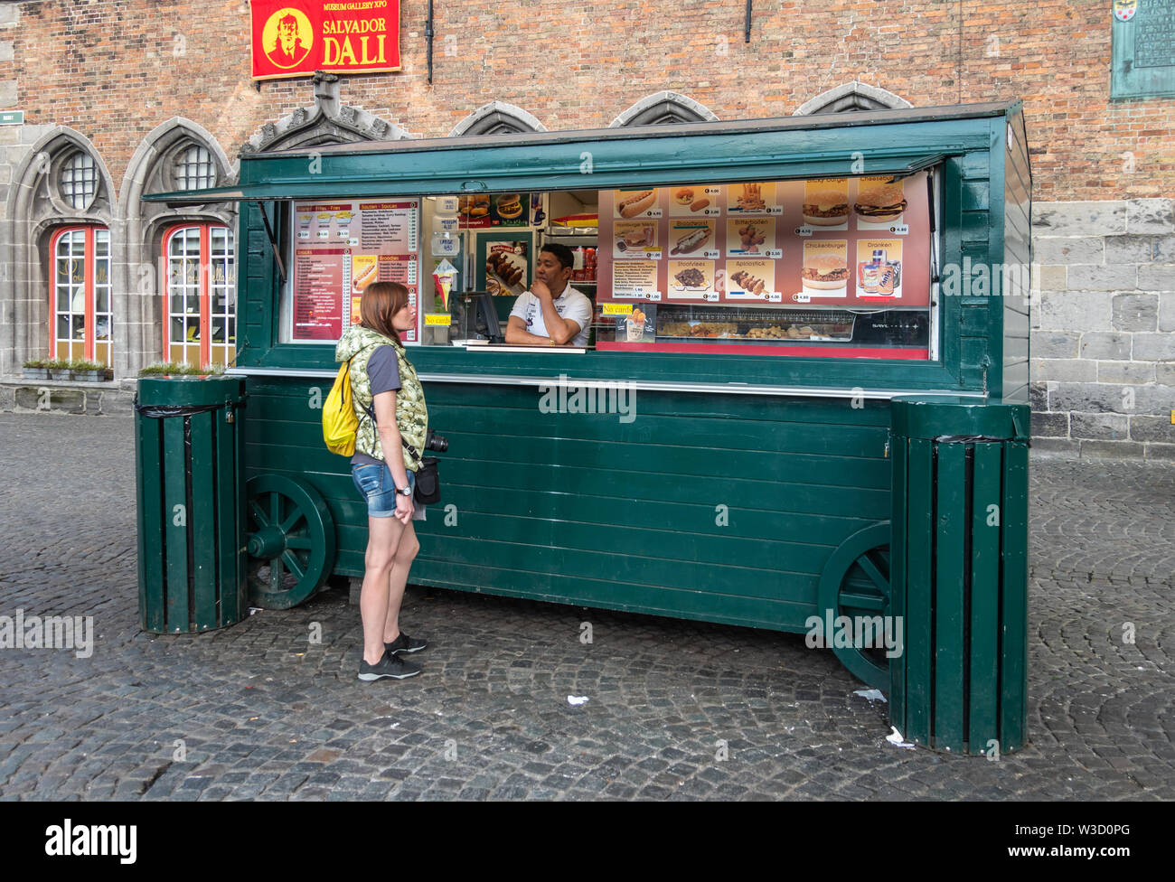 Bruges, Flanders, Belgium -  June 15, 2019: Green wooden inconic French Fries booth in front of Belfry. Vendor and customer. Red banner for Dali exhib Stock Photo