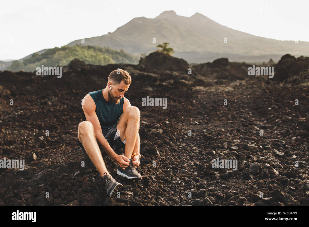 Young man sitting and tying running shoes before trail running outdoors. Volcanic landscape and mountain view on background. Stock Photo