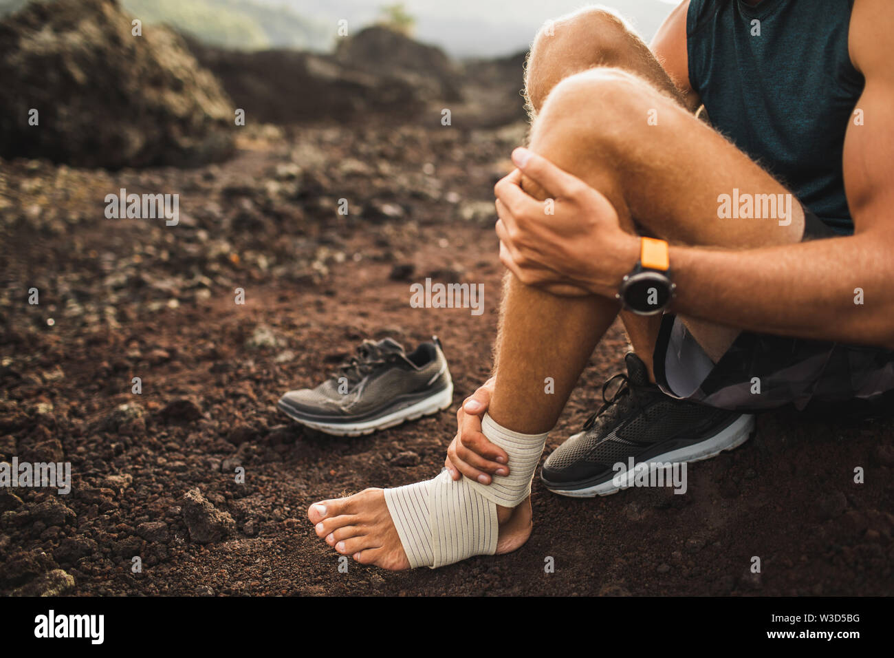 Man bandaging injured ankle. Injury leg while running outdoors. First aid for sprained ligament or tendon. Close-up on dark background. Stock Photo