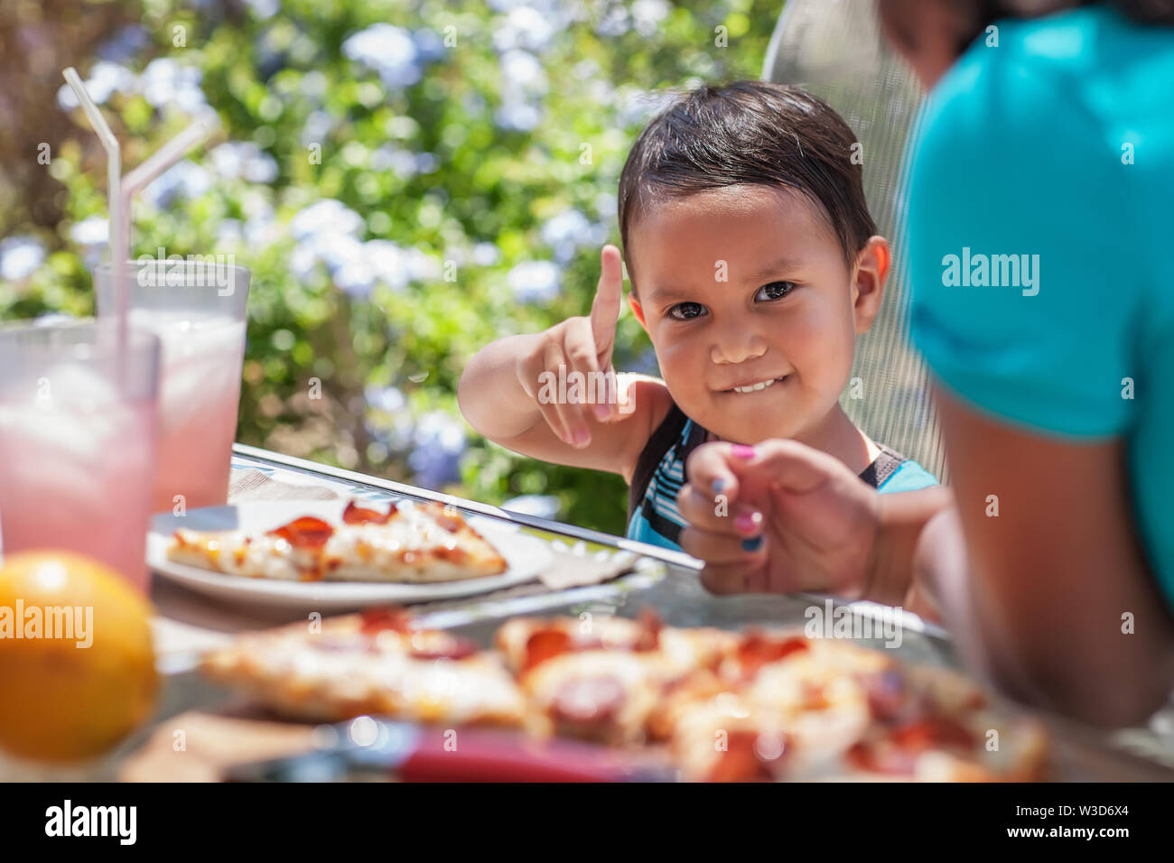 Young brother loves eating cheese pizza for lunch in an outdoor backyard patio with his older sister, he gestures number one with finger. Stock Photo