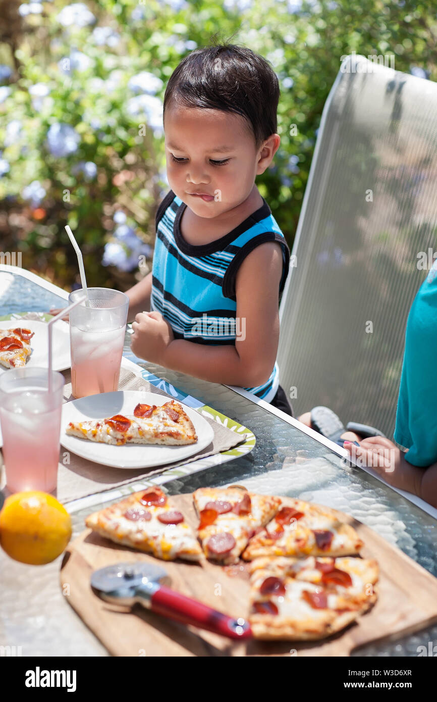 A little kid bites his lip as he looks at his lunch meal consisting of turkey pepperoni pizza and pink lemonade. Stock Photo