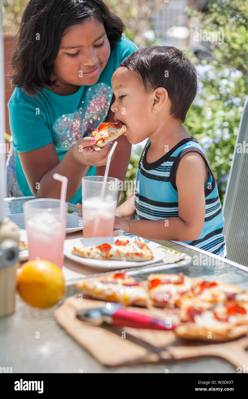 Siblings enjoy an outdoor lunch, while the big sister feeds her little brother a slice of pizza. Stock Photo