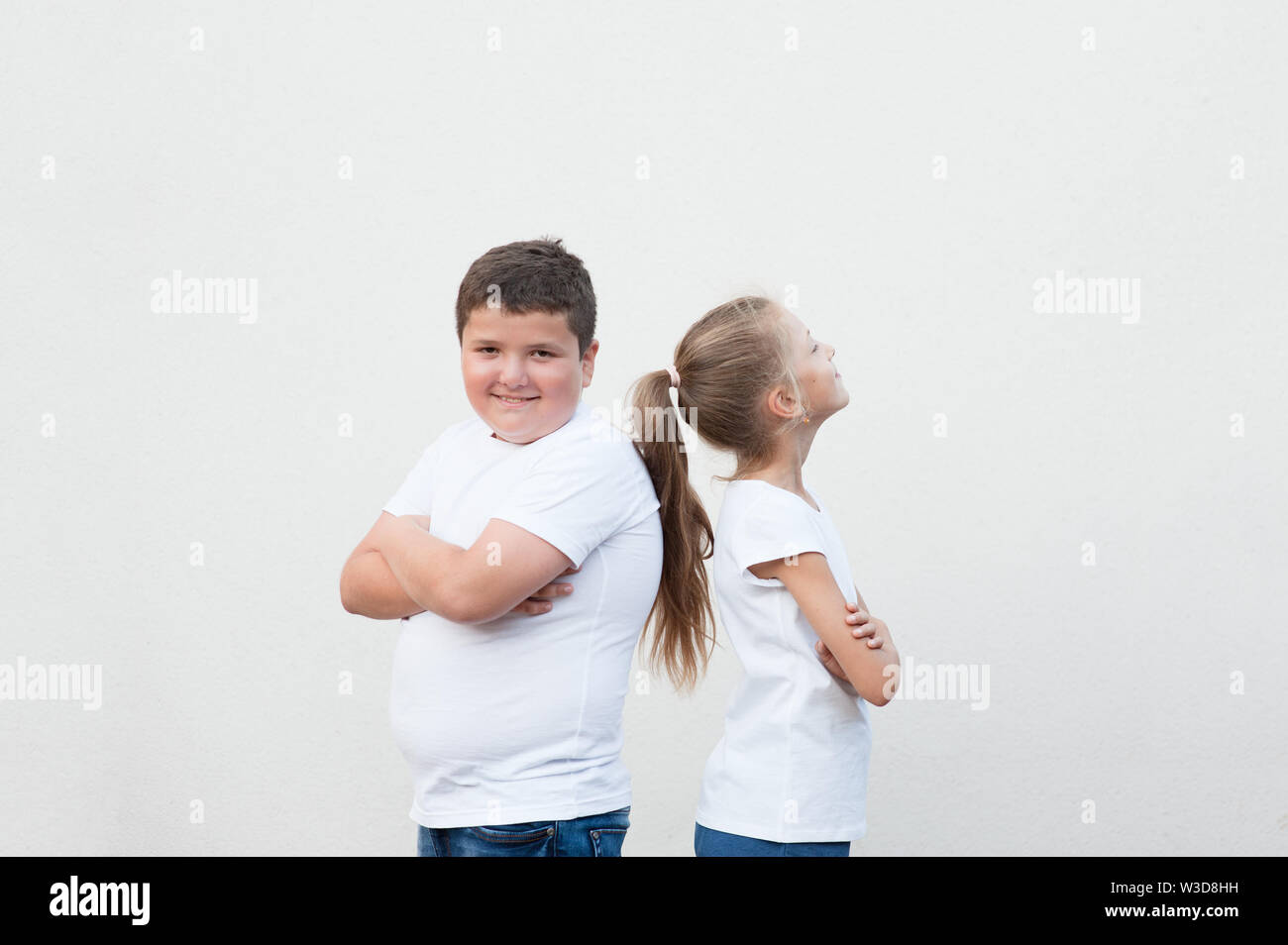concept of thin girl and thick boy in white shirt standing back to back on white wall background Stock Photo