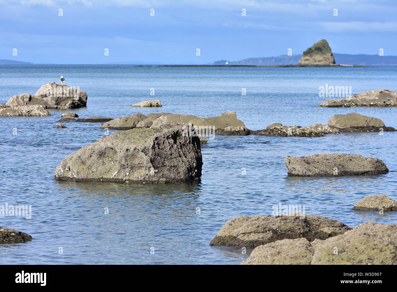 Rocks protruding from calm sea surface with islet and rocky reef in background. Stock Photo