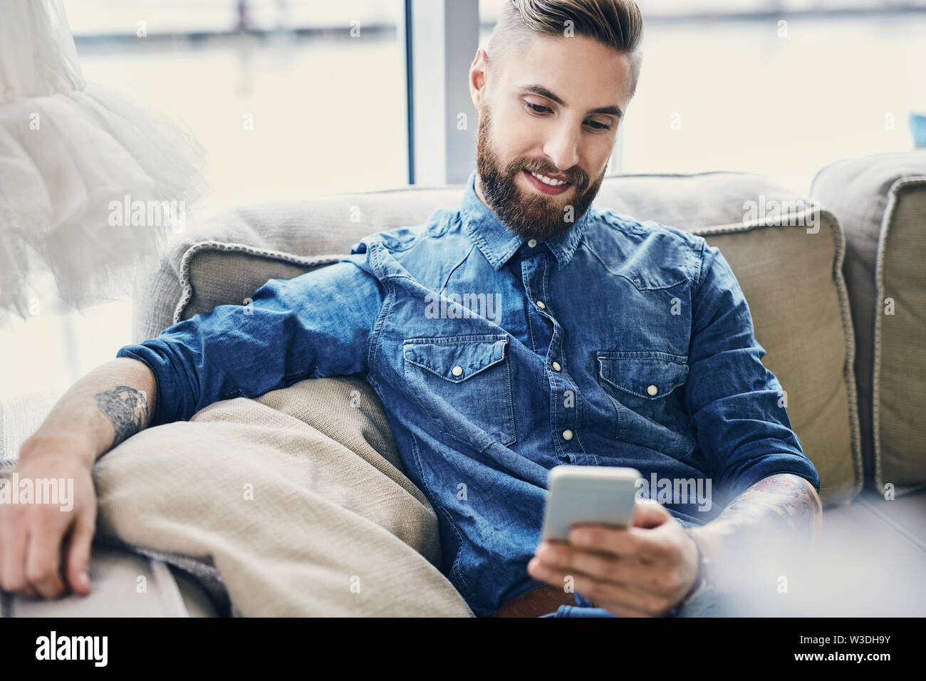 Happy tattooed man using his smartphone sitting sofa Stock Photo