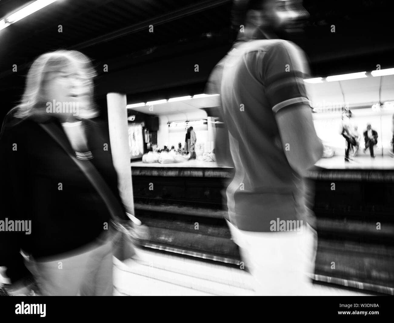 Defocused blur people silhouettes walking inside Barcelona Metro station with silhouettes of homeless migrants sleeping on the train platform - black and white. Stock Photo