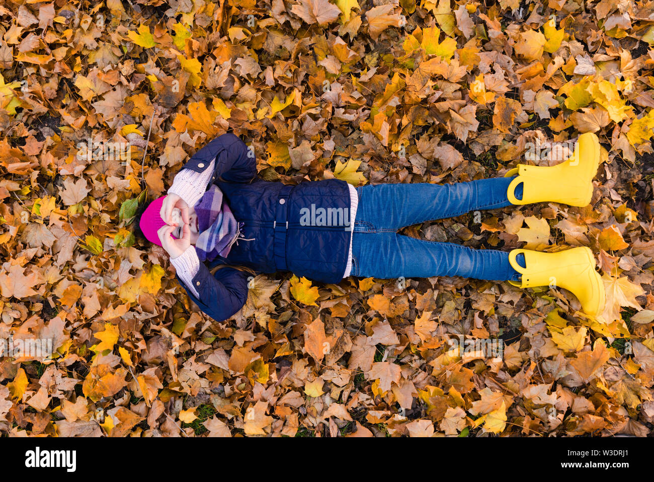 Lovely kid at autumn park on foliage up view. Girl lay at leaves and show heart. View from above. Stock Photo