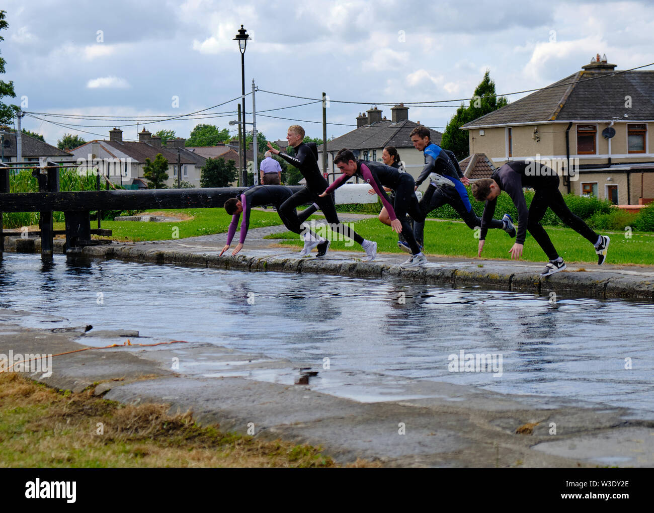 Line of teenagers jumping off in Grand Canal wearing wet suits by Suir Road bridge on hot summer day in Dublin, Ireland Stock Photo