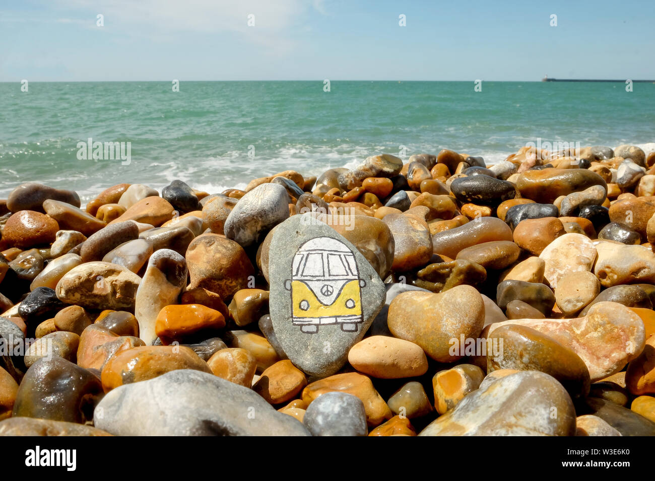 A yellow and white camper van painted onto a pebble on a pebble, beyond the pebbles is the blue sea and blue sky Stock Photo