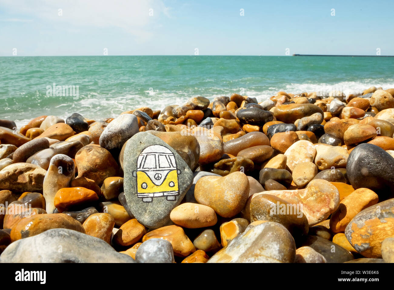 A yellow and white camper van painted onto a pebble on a pebble, beyond the pebbles is the blue sea and blue sky Stock Photo