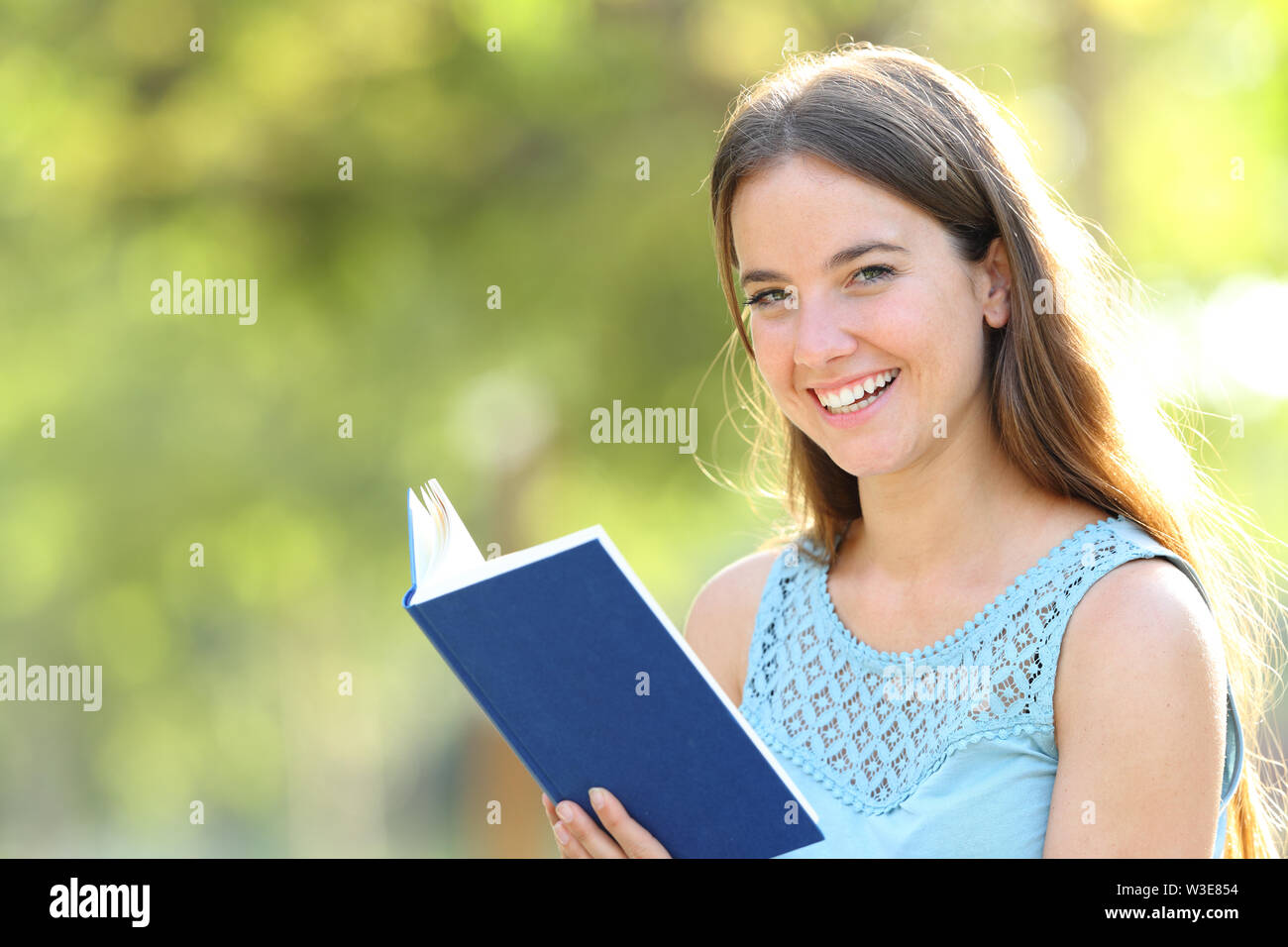Happy woman looks at camera holding a blank paper book on green background in a park Stock Photo