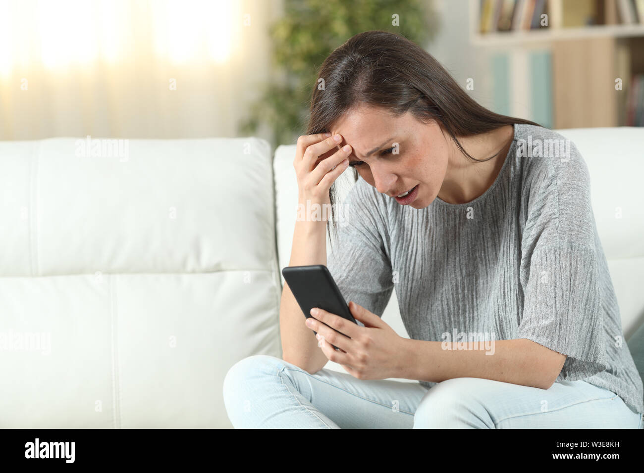 Worried woman checking smart phone sitting on a couch in the living room at home Stock Photo