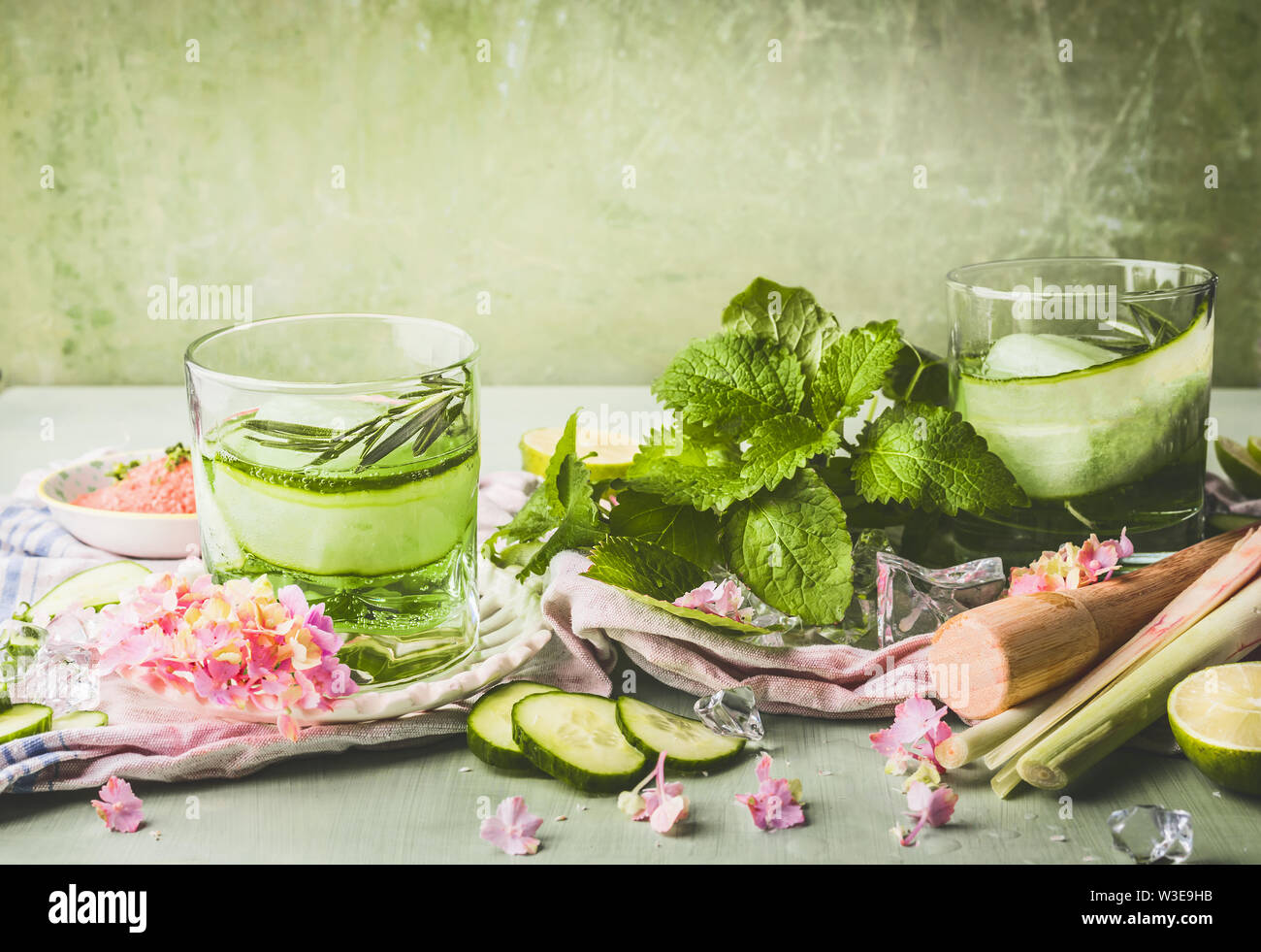 Classes with cucumber lemonade or detox infused water and ice cubes on table with ingredients. Green color summer refreshing drinks preparation Stock Photo