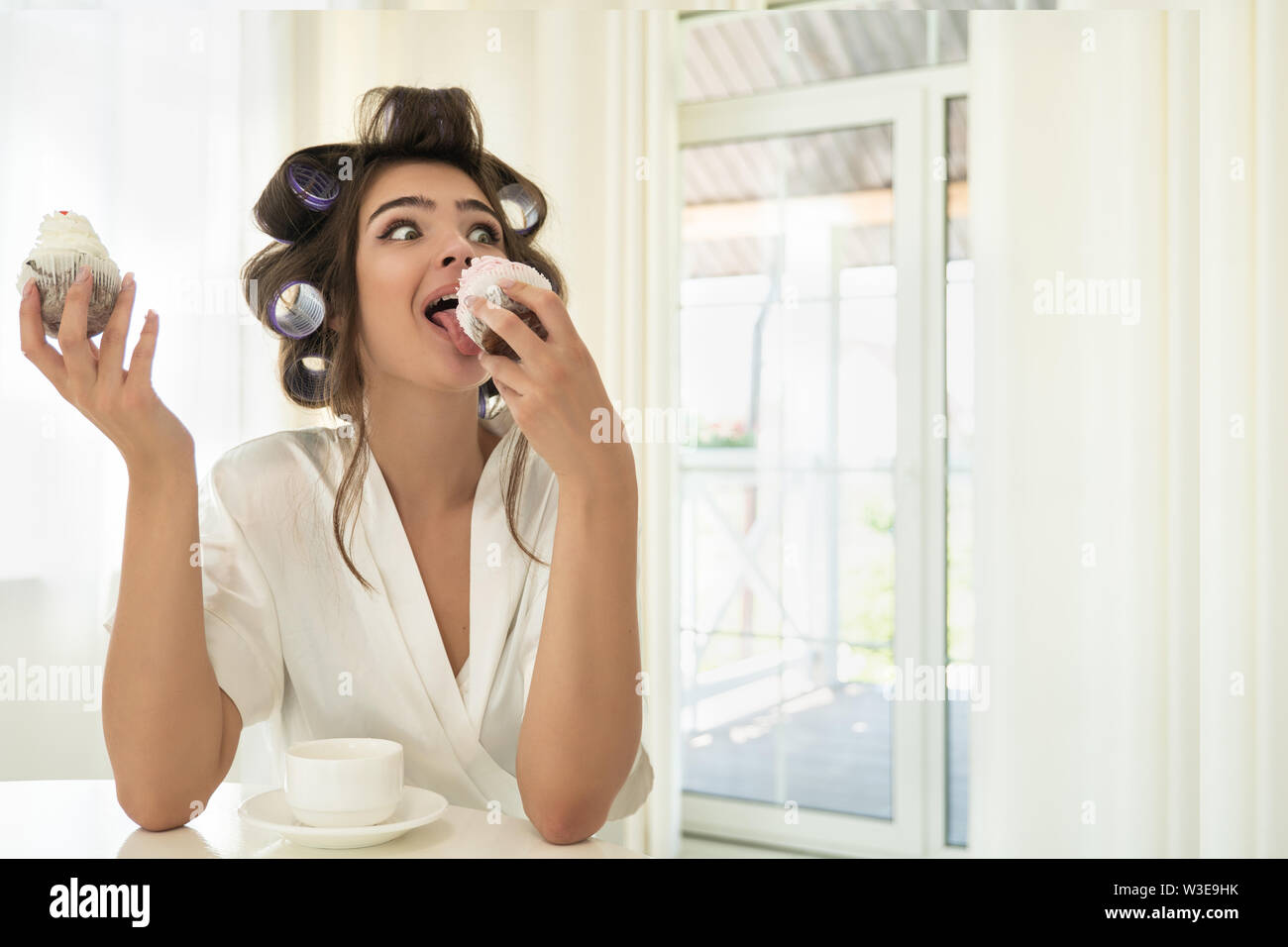 beautiful funny young brunette woman in hair curlers licking one cupcake and holding another in her hand sitting with cup of coffee in bright kitchen Stock Photo