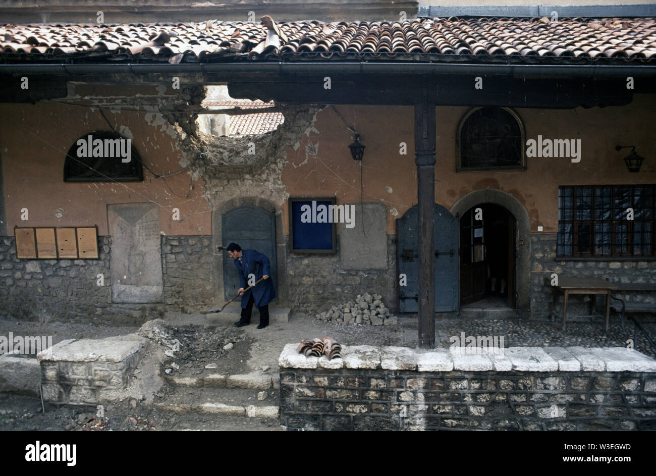 5th April 1993 During the Siege of Sarajevo: in the courtyard of the oldest Orthodox church in Sarajevo, a man shovels up debris below a large hole in the wall made by a mortar bomb or artillery shell. Stock Photo