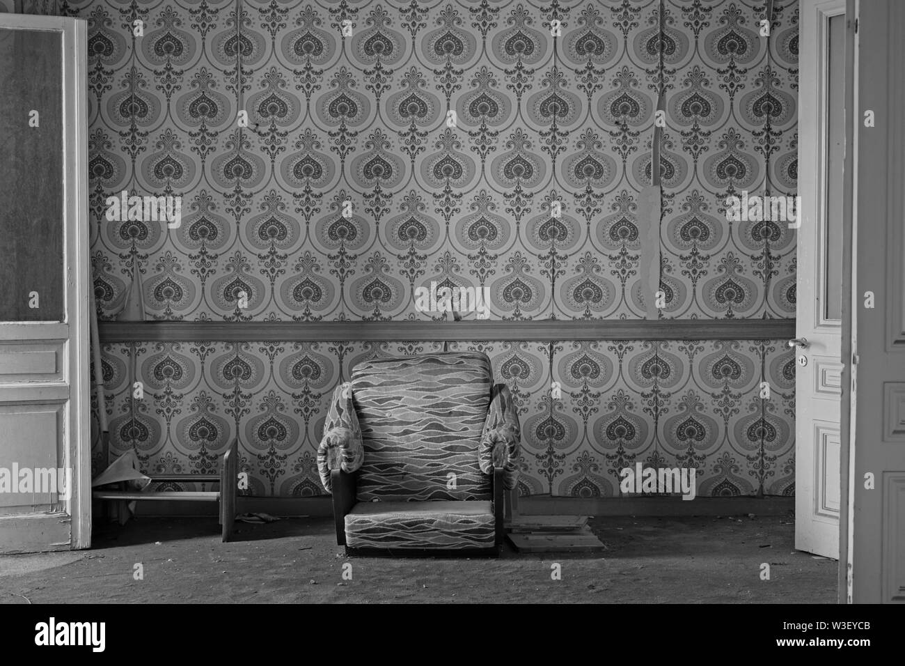 Broken armchair on dirty carpet and torn wallpaper on living room wall of an abandoned house. Black and white. Stock Photo