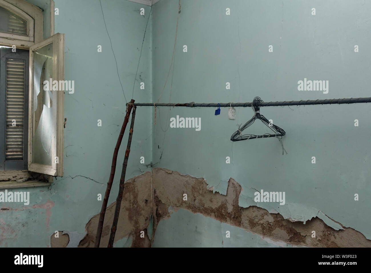 Broken window peeling wall and makeshift clothesline with rusty hangers in decayed blue room interior. Stock Photo