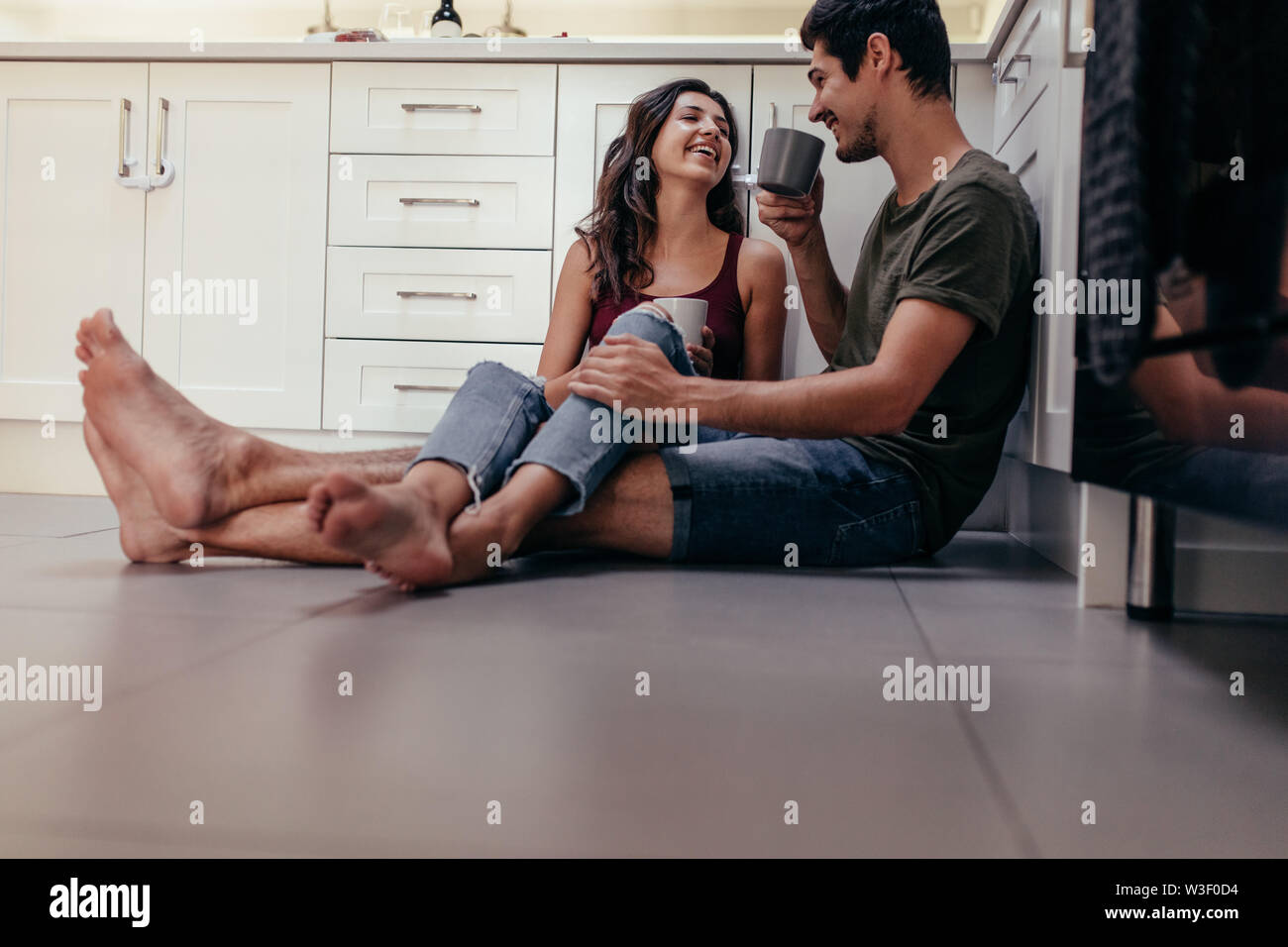 Happy young couple relaxing in kitchen having coffee. Young man and woman sitting on kitchen floor drinking coffee Stock Photo