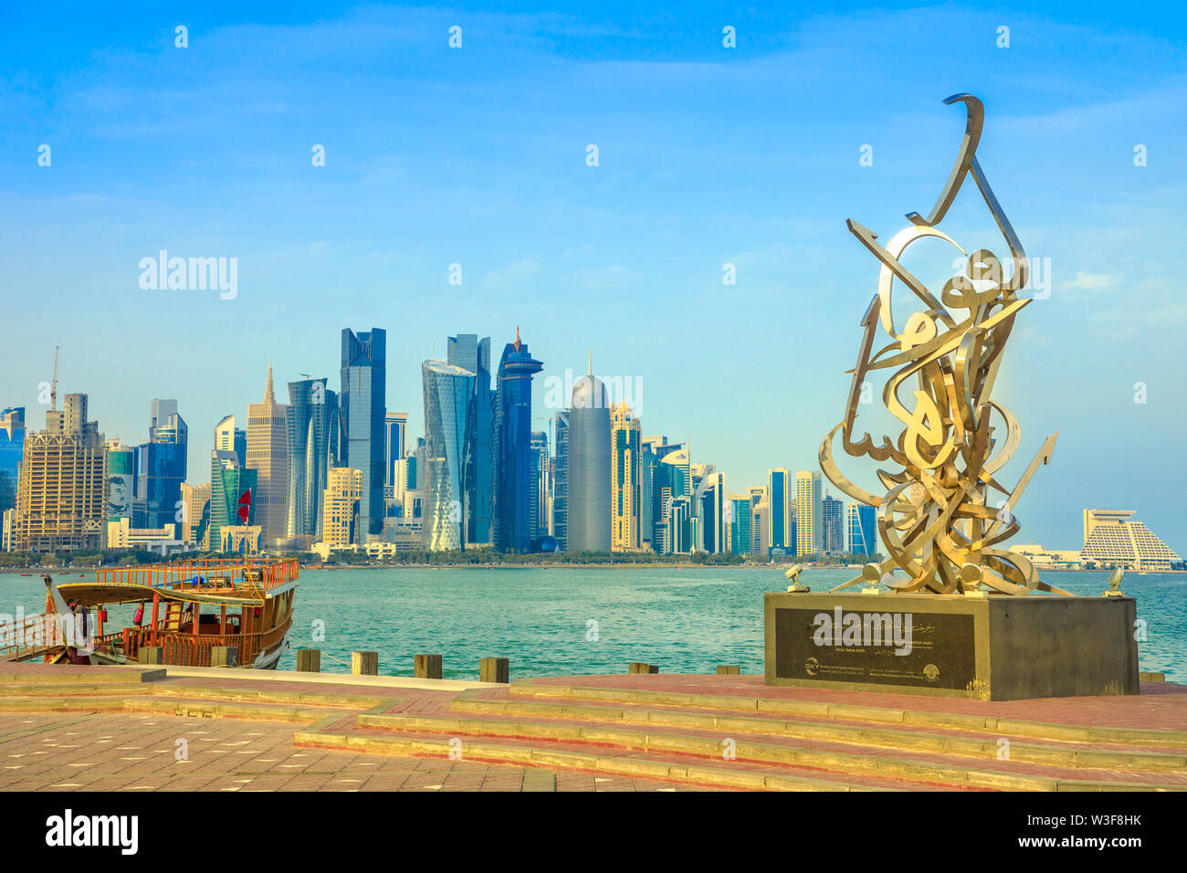 Doha, Qatar - February 23, 2019: Calligraphy sculpture on the corniche seaside promenade, wooden dhow and Doha West Bay skyline with Doha Tower, Salam Stock Photo