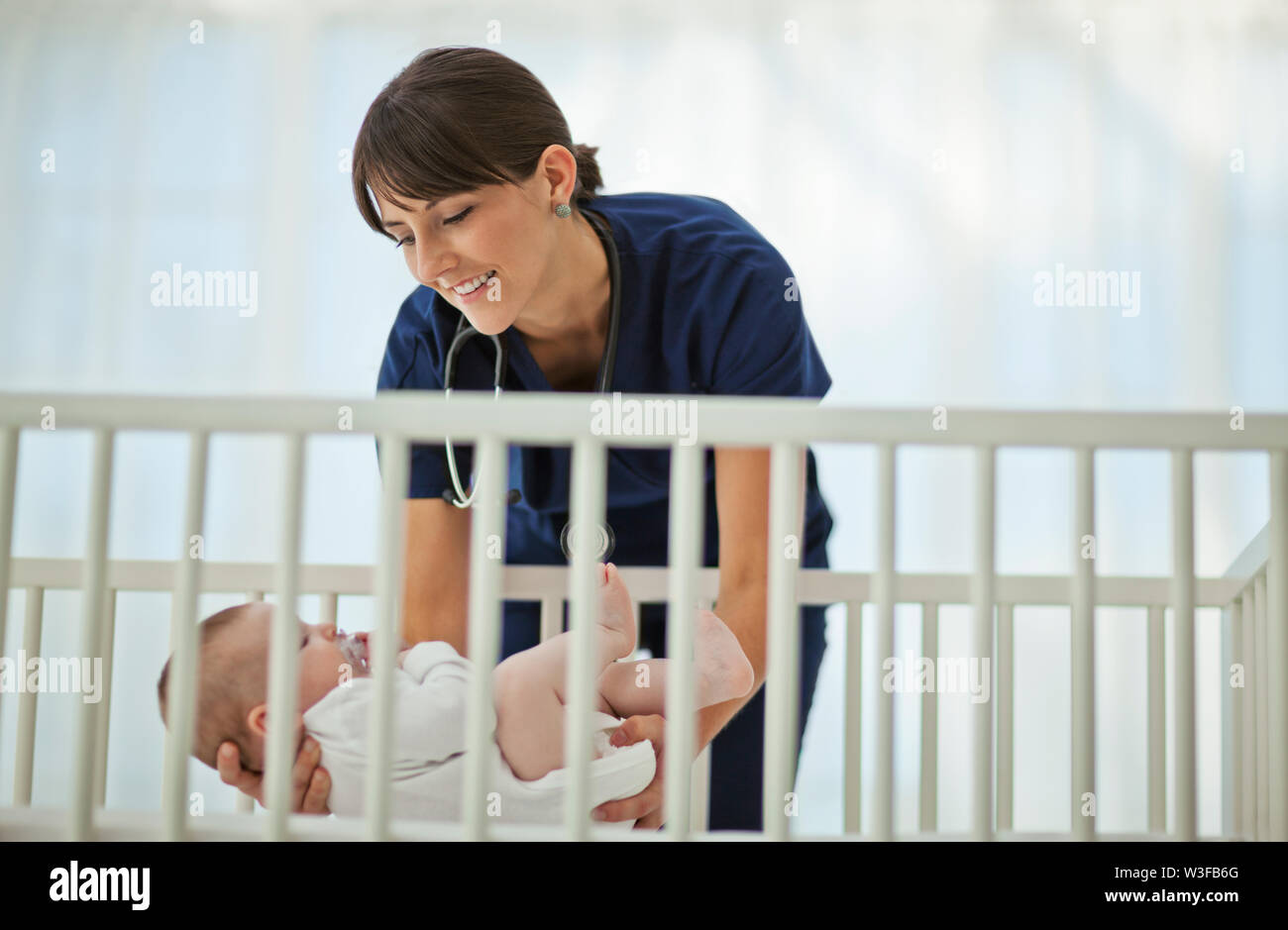 Smiling young nurse gently laying a sleepy baby down in his crib. Stock Photo