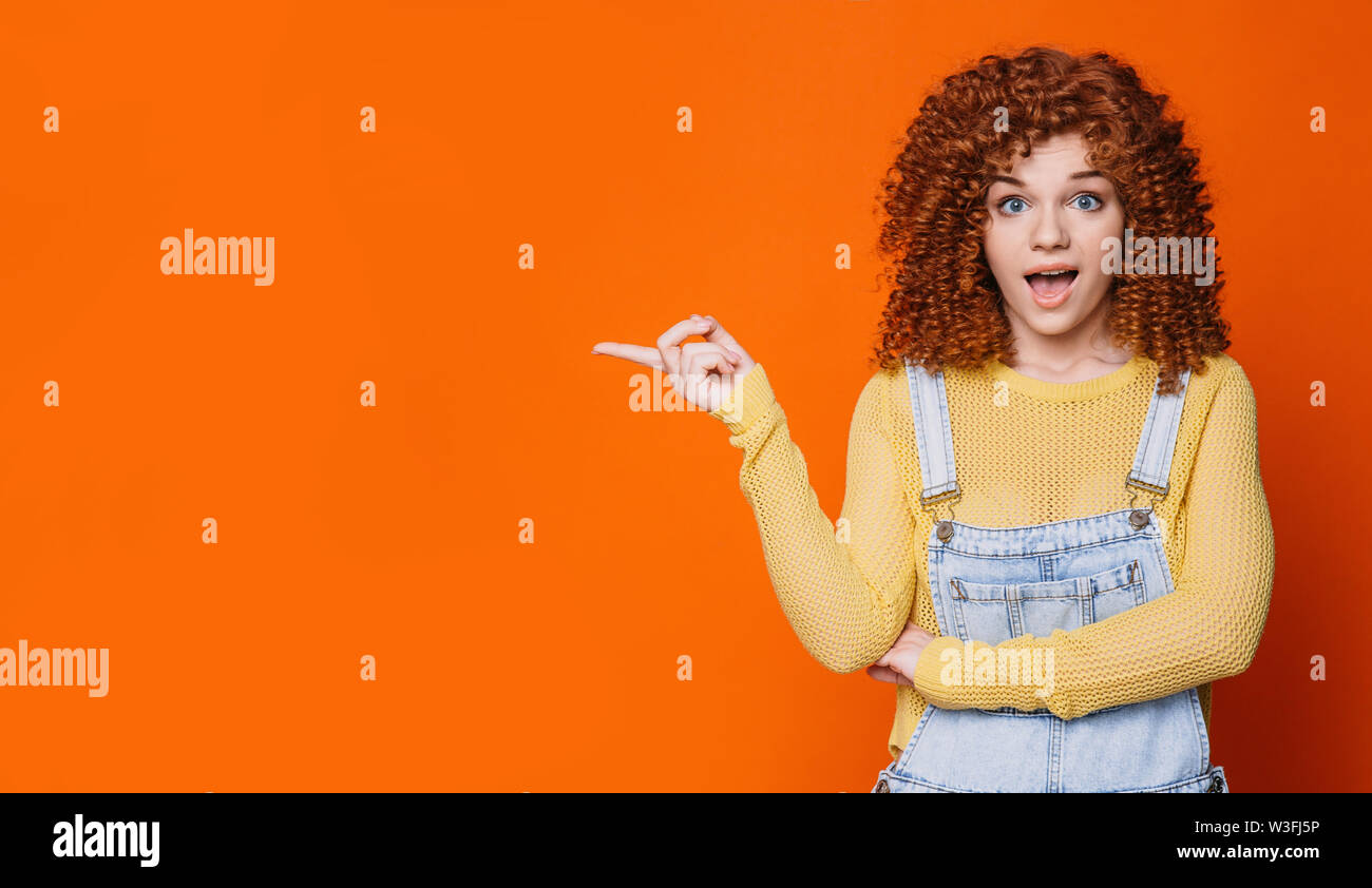 Wow emotion , curly haired woman advertising product, pointing hand on empty orange wall. Stock Photo
